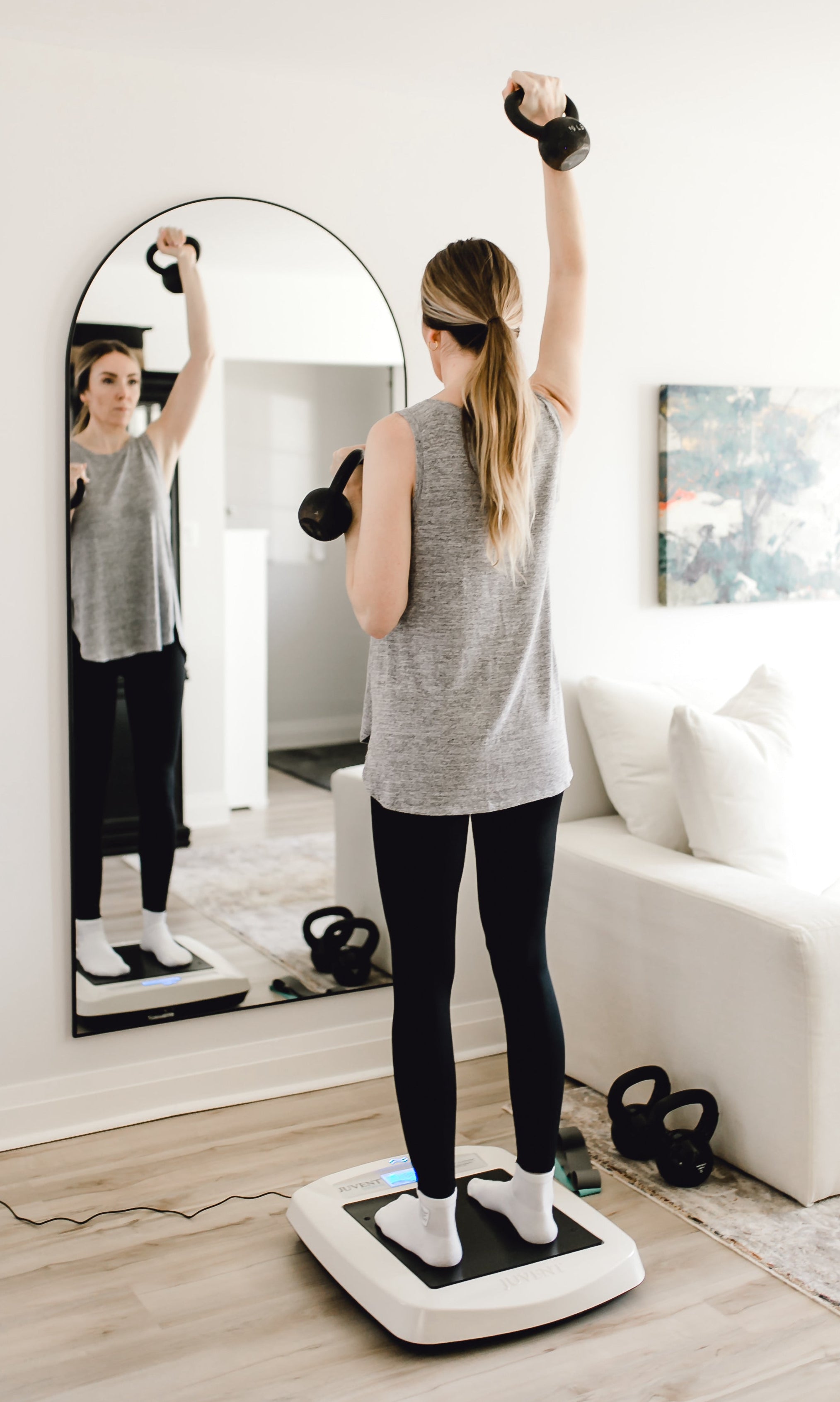 Person using a vibration plate in a home setting with a mirror and fitness equipment.